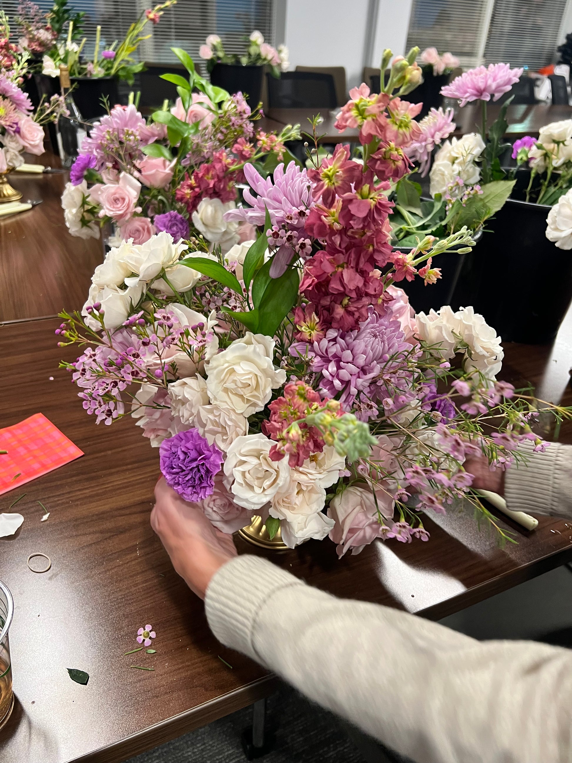 A person arranging a floral composition with a variety of flowers including shades of pink, white, and green, at a table.
