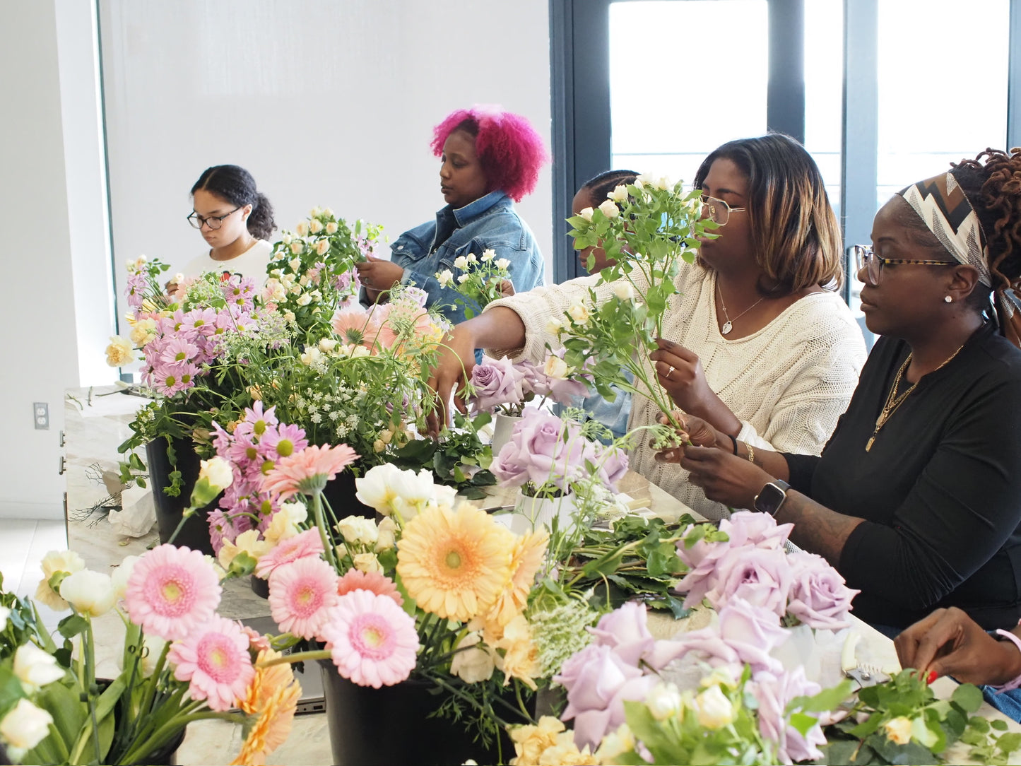 A group of people participating in a floral design workshop, arranged with individuals behind various tables, each creating a floral arrangement with a variety of flowers.