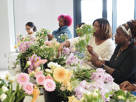A group of people participating in a floral design workshop, arranged with individuals behind various tables, each creating a floral arrangement with a variety of flowers.